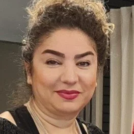 Close-up van een vrouw met krullend haar en een glimlach, dragend zwarte kleding en een lichte achtergrond.