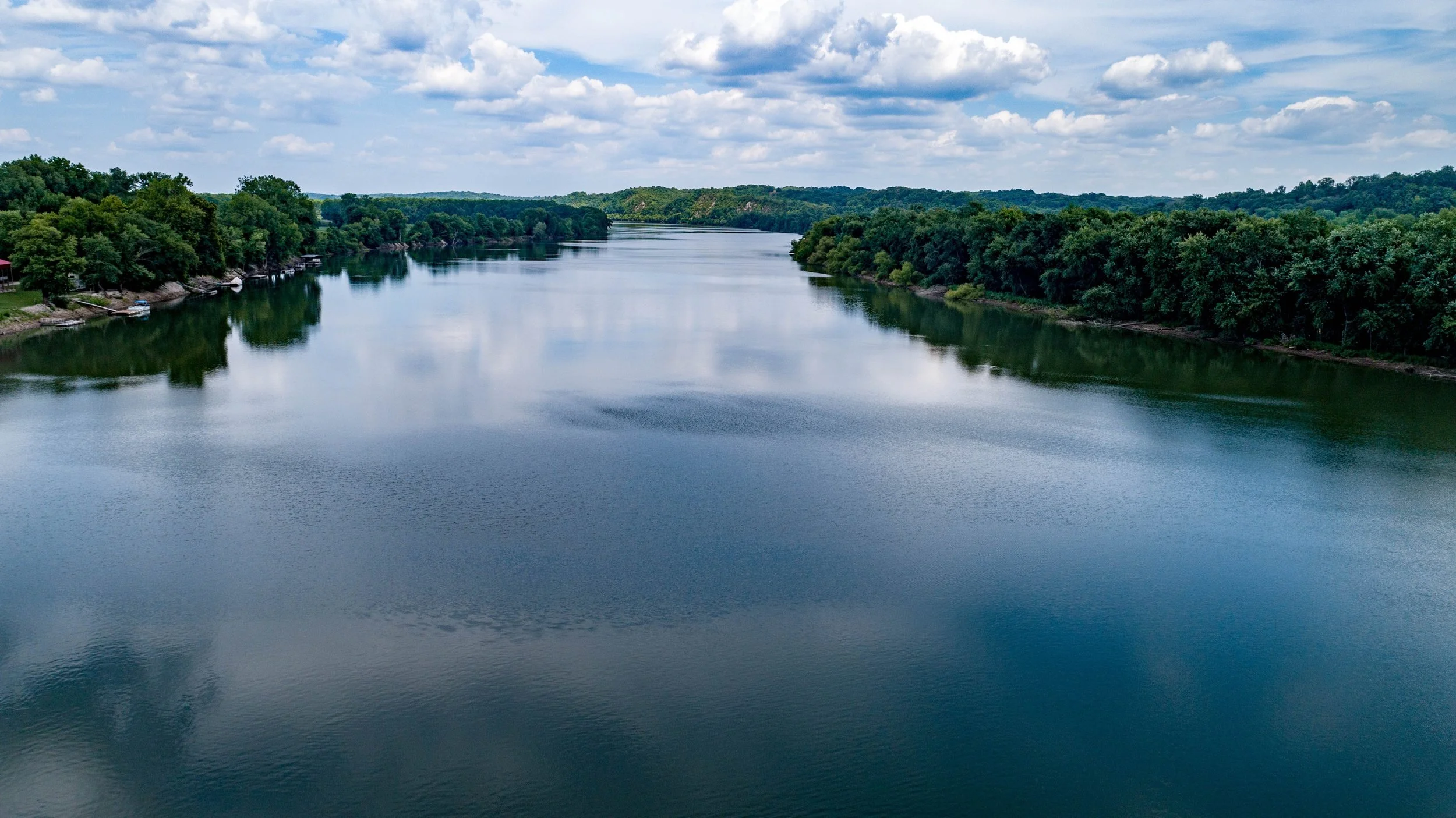 A wide view of a calm river surrounded by lush green trees under a partly cloudy sky.