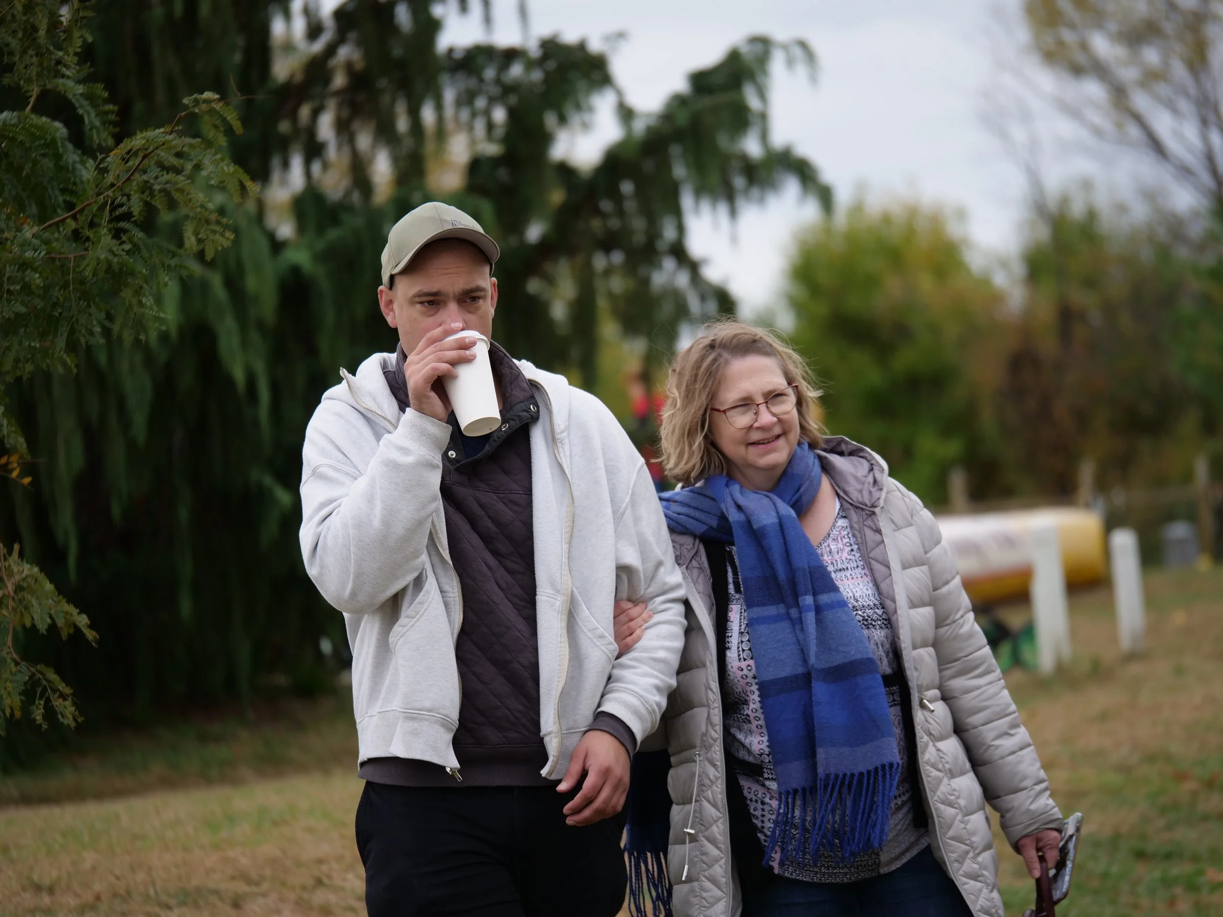 Noah Worcester, wearing a beige cap, white jacket, and dark shirt, drinks coffee from a cup while walking outdoors with his mother, Rachel, who is wearing glasses, a light puffer jacket, and a blue scarf.