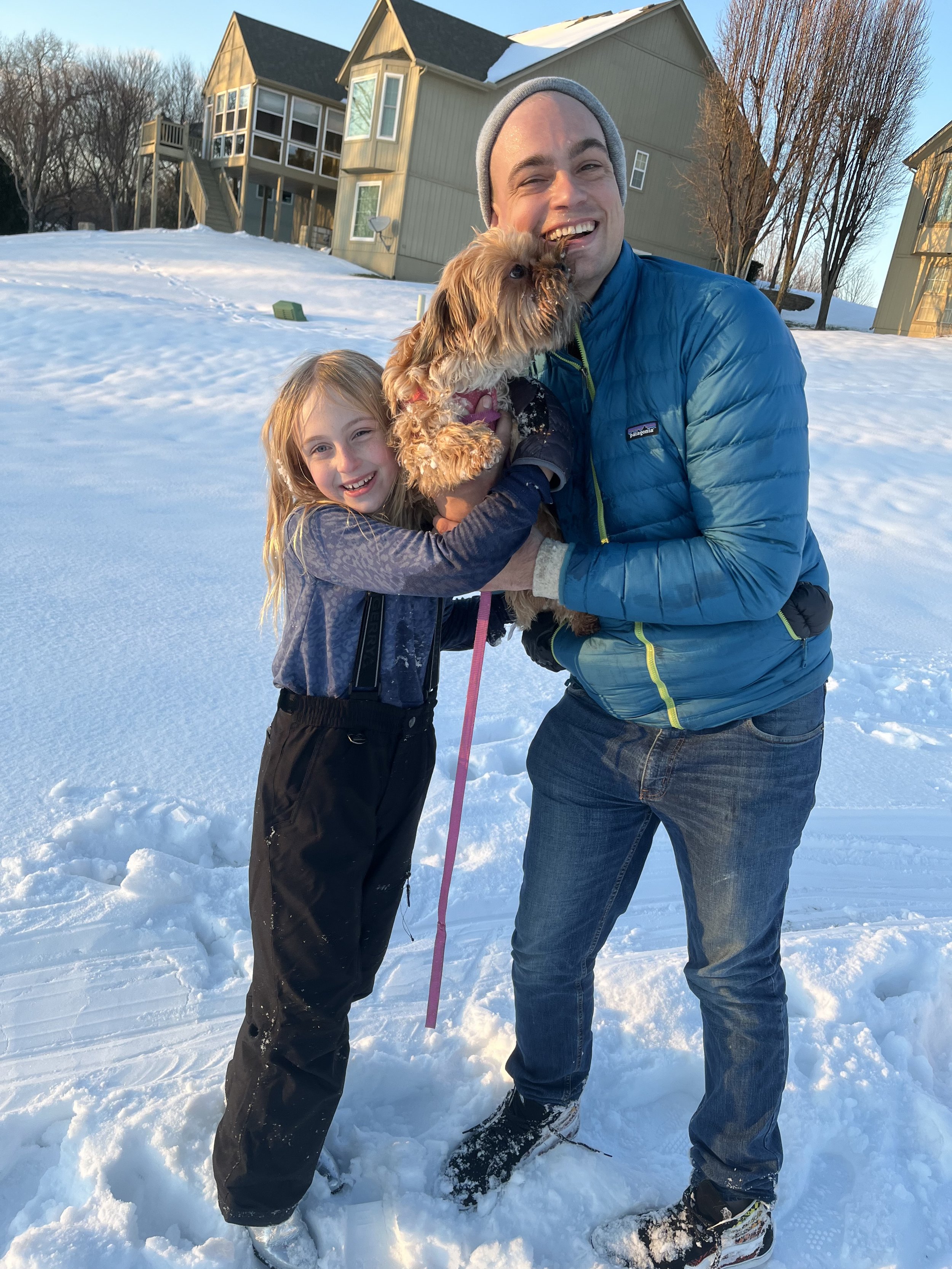 Noah Worcester and his daughter, Zoe, smile while standing in the snow outside a row of houses, holding their small dog, Pixie, during a winter day.
