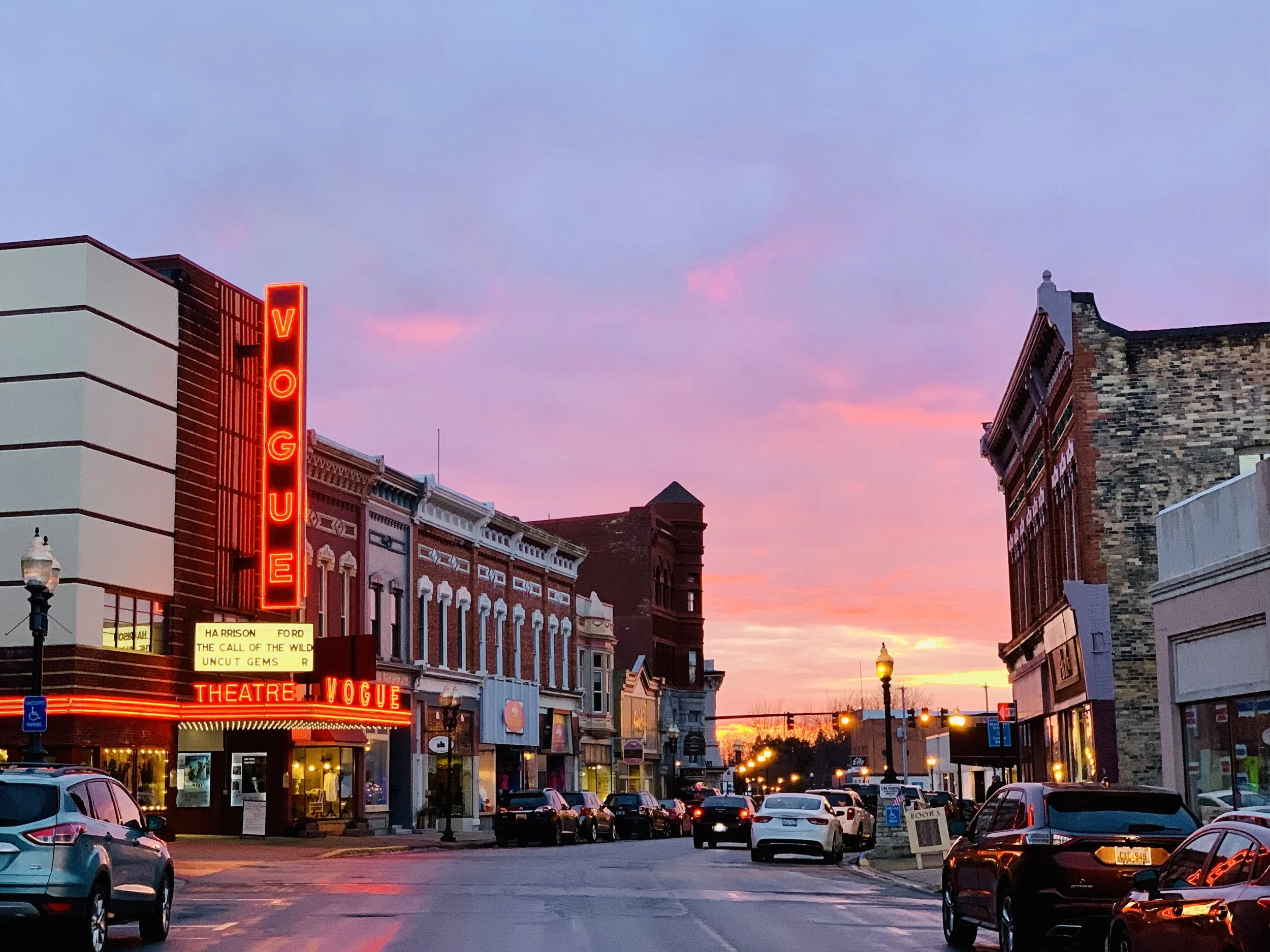 A lively photo of downtown River Street in Manistee, MI at sunset with boutique shops and theaters, including the Vogue Theatre with a neon sign, with parked cars along the street and a colorful sky.