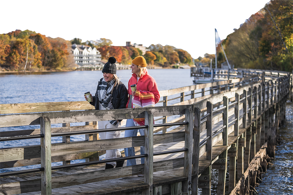 Two women walking on a wooden pier beside the Manistee River, smiling and holding coffee cups, in a fall setting with colorful trees and residential buildings in the background.