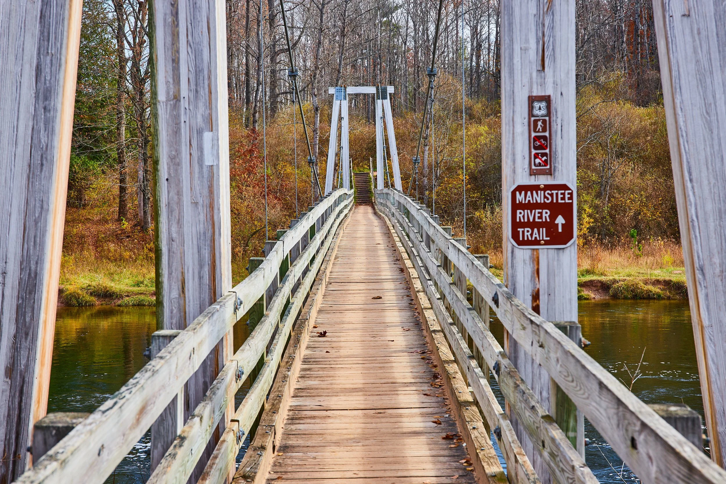 Wooden footbridge over a river with a sign indicating the Manistee River Trail, surrounded by trees with autumn foliage.