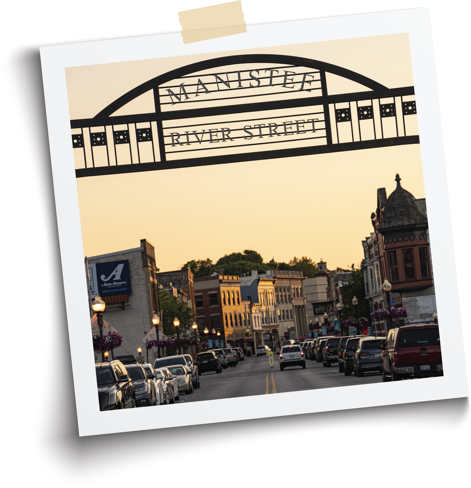 The photo shows a street scene on Manistee River Street in a historic downtown area, with buildings on both sides, streetlights, flowers, and parked cars, under a light sky at sunset.