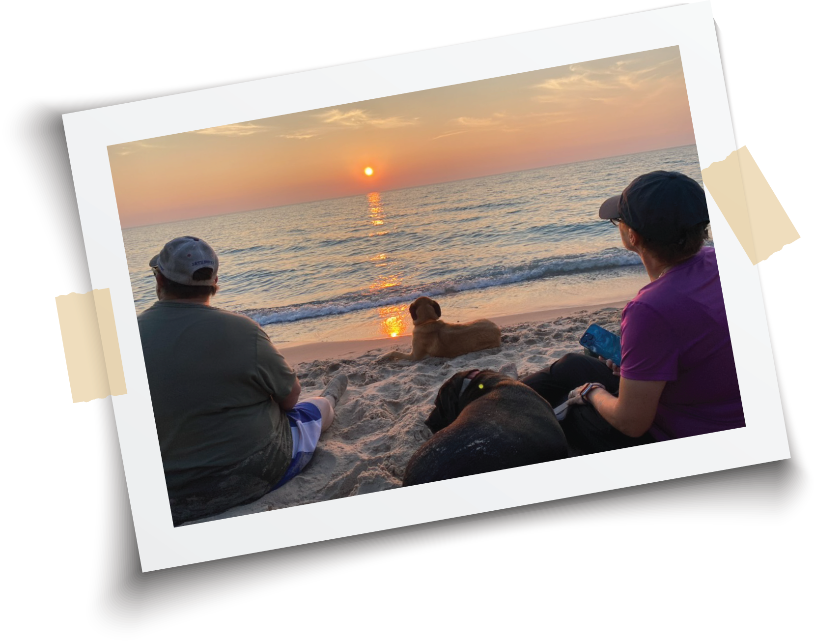 Two people and two dogs are sitting on the beach of Lake Michigan during sunset, facing the ocean with a calm sea and a partly cloudy sky.