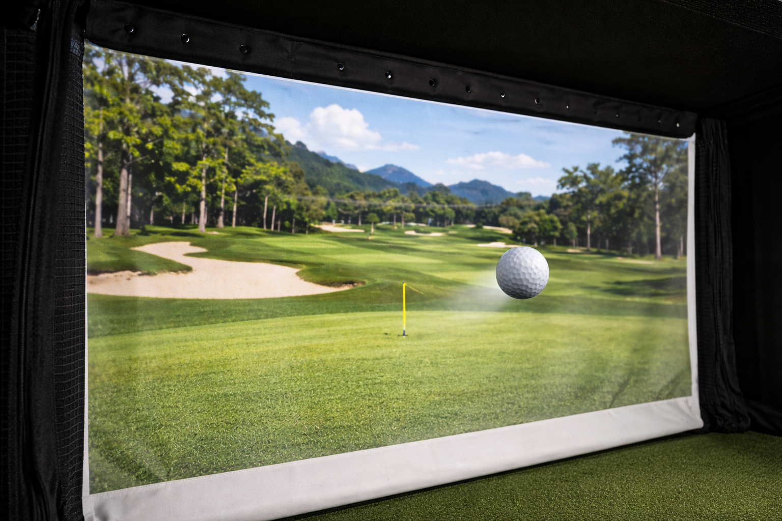 Indoor golf simulation showing a virtual golf course, with a golf ball in mid-air near the green and a yellow flag in the distance, surrounded by trees and mountains under a partly cloudy sky.