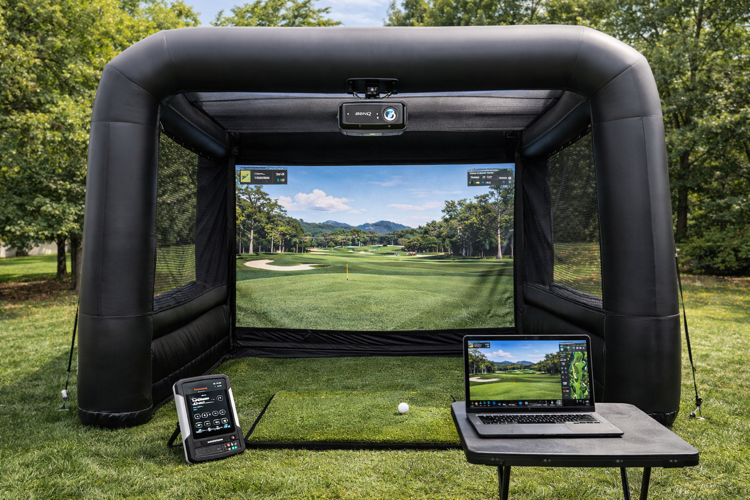 Golf simulator setup with a large screen displaying a virtual golf course, a laptop on a small table showing the same course, and control devices in an outdoor setting with trees and grass.