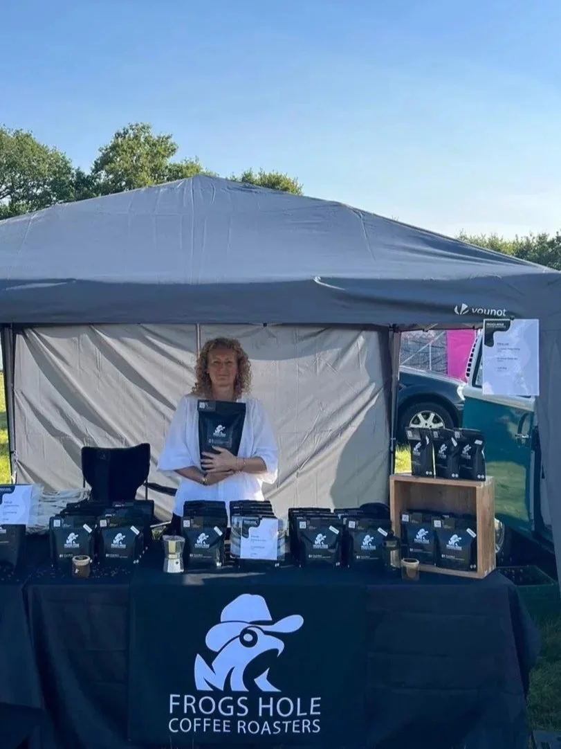 A woman standing behind a table at a booth for Frogs Hole Coffee Roasters, with coffee bags displayed on the table, under a tent on a sunny day with trees and a car in the background.