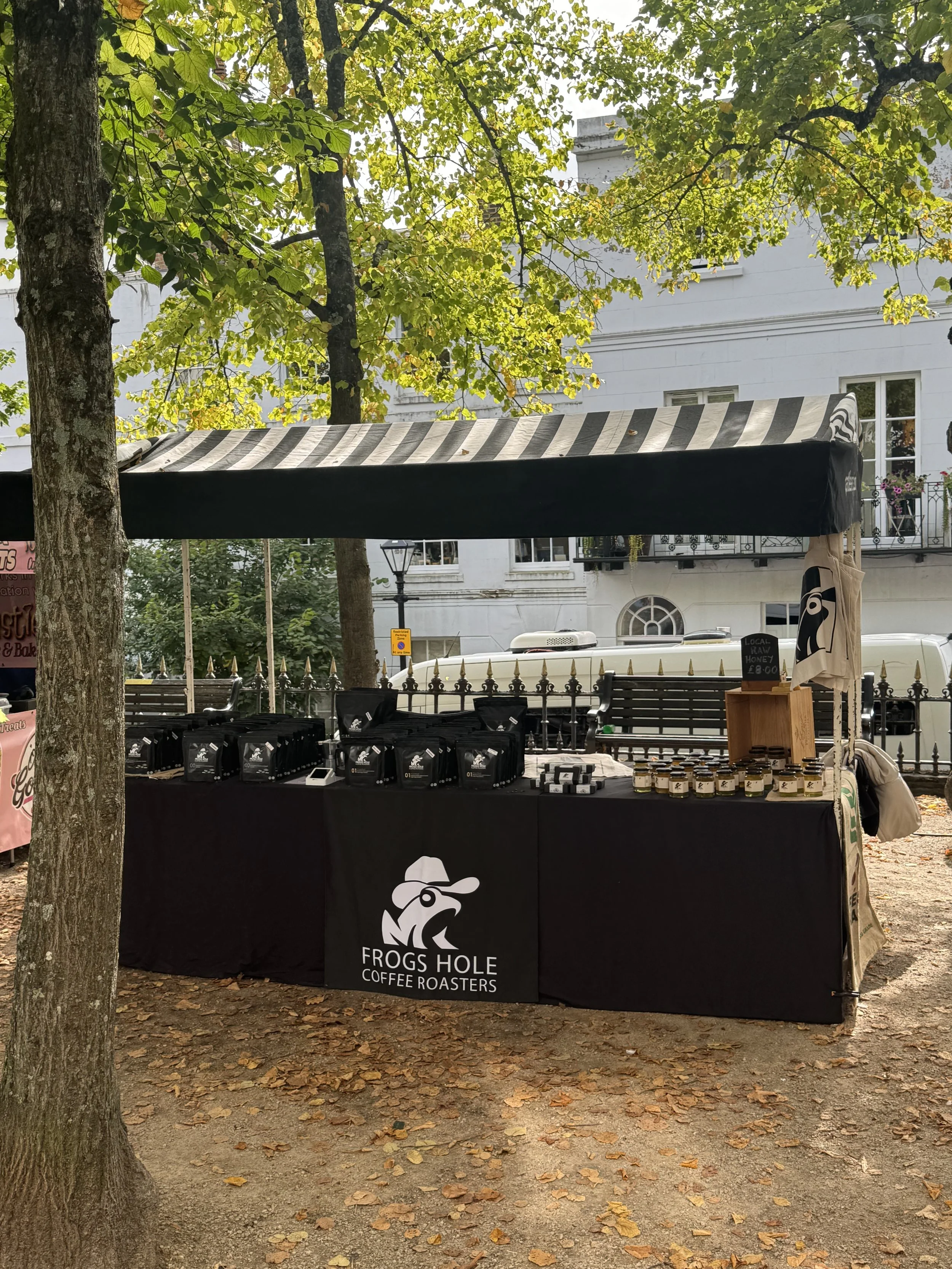Outdoor coffee stand with a black tablecloth featuring a frog logo and the text 'Frogs Hole Coffee Roasters,' set under a canopy with coffee bags and jars on display, trees with green leaves in the background.