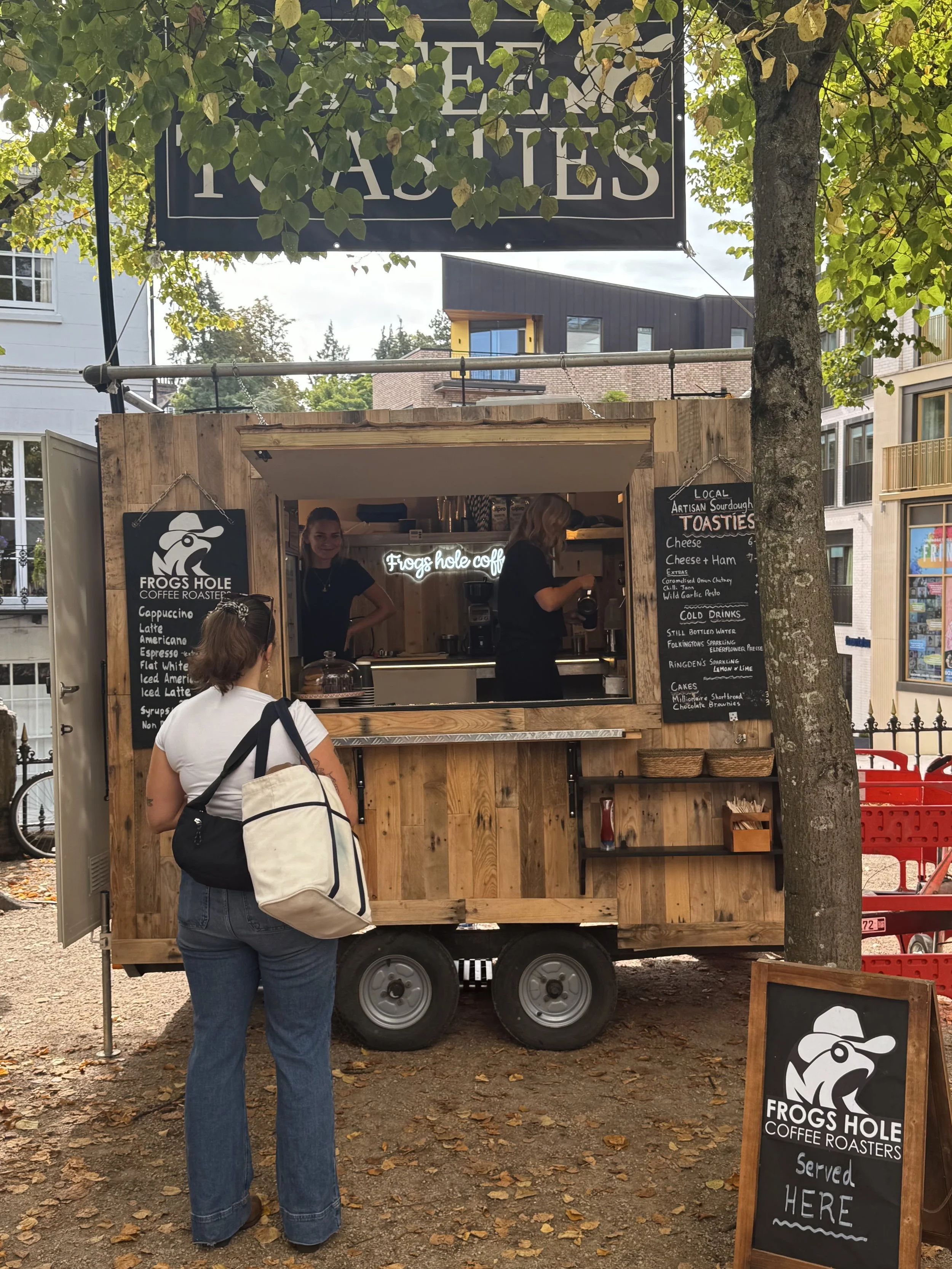 A woman with a large bag waiting at a small wooden food cart named Frogs Hole Coffee Roasters with a menu board outside on a sidewalk with fallen leaves and a tree.