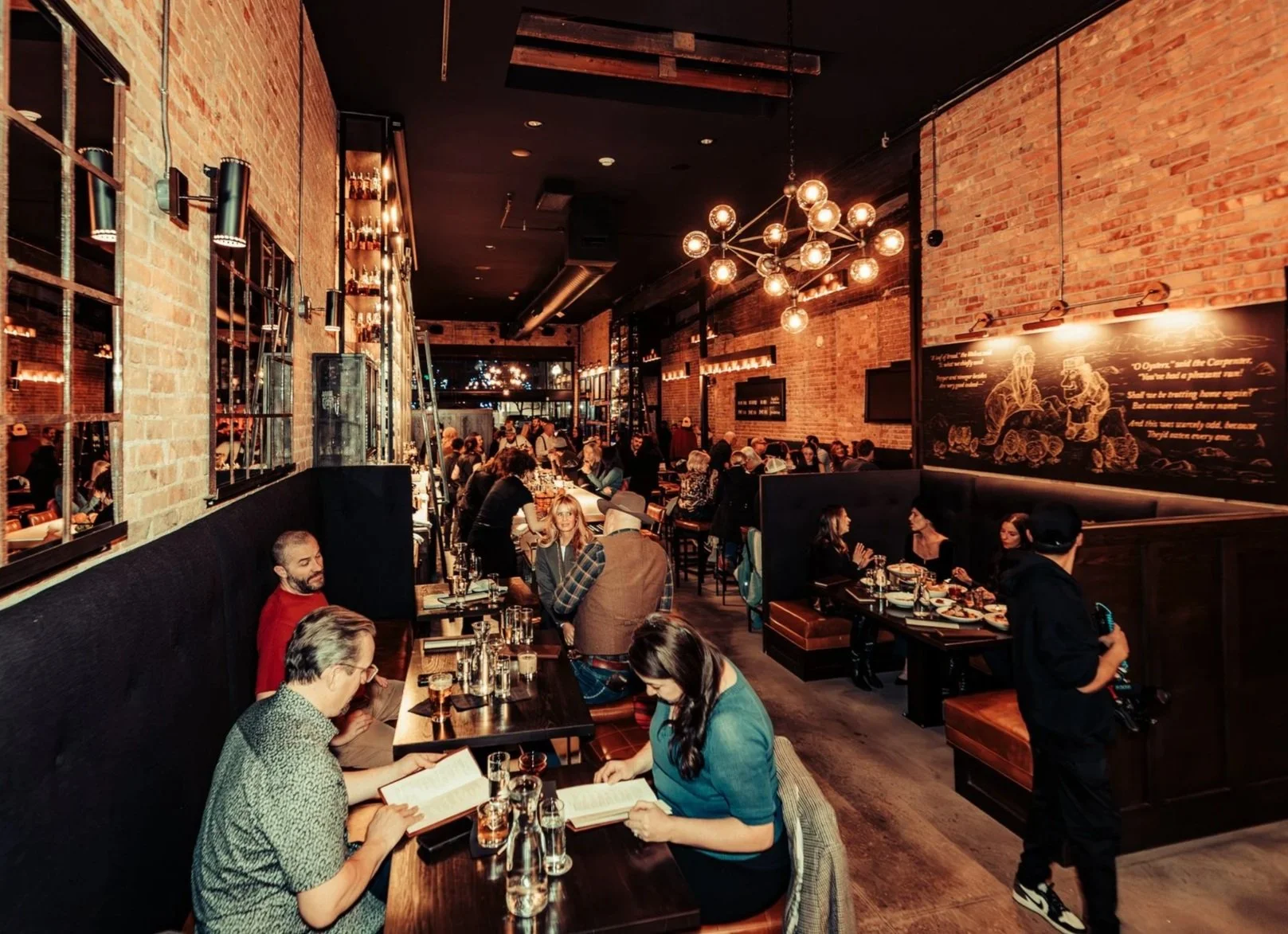 A lively restaurant with exposed brick walls, black ceiling, and modern chandelier lights, filled with people dining and socializing.
