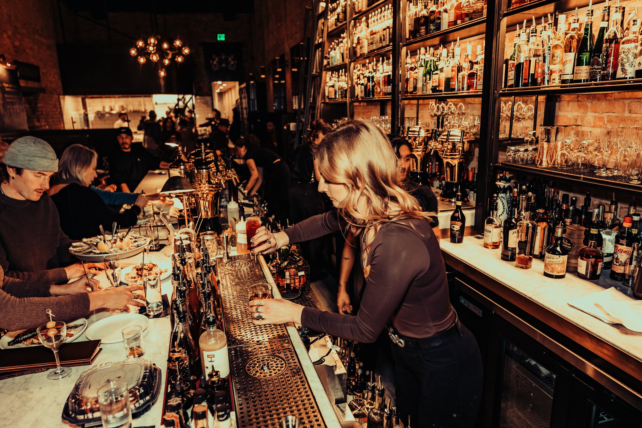 A bartender pouring a drink at a busy bar with bar patrons sitting at the counter, and shelves of bottles and glasses behind the bar.