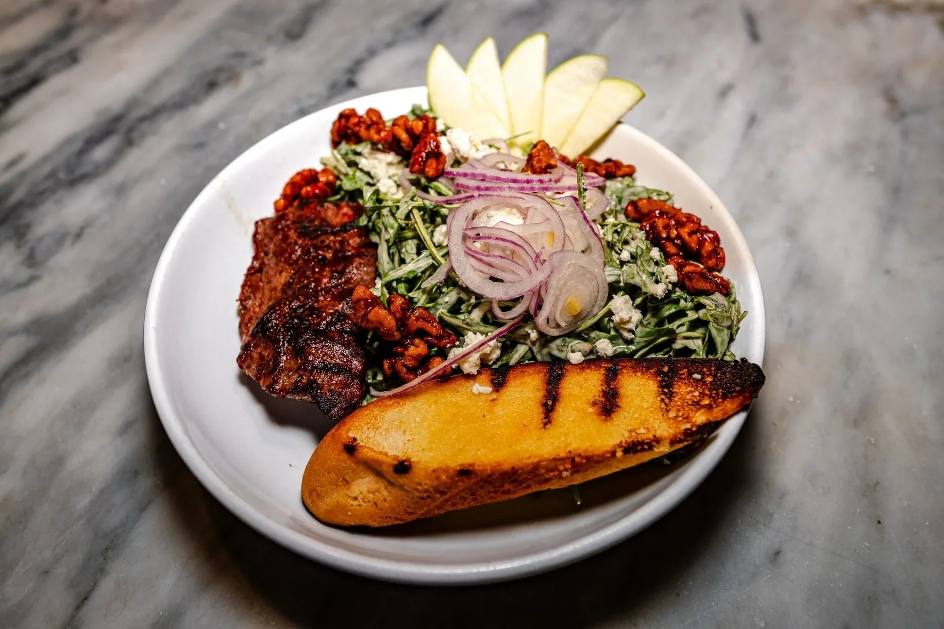 A white bowl containing a grilled bread slice, a piece of grilled meat, a salad with sliced red onions, crumbled cheese, walnuts, and apple slices on a gray marble surface.