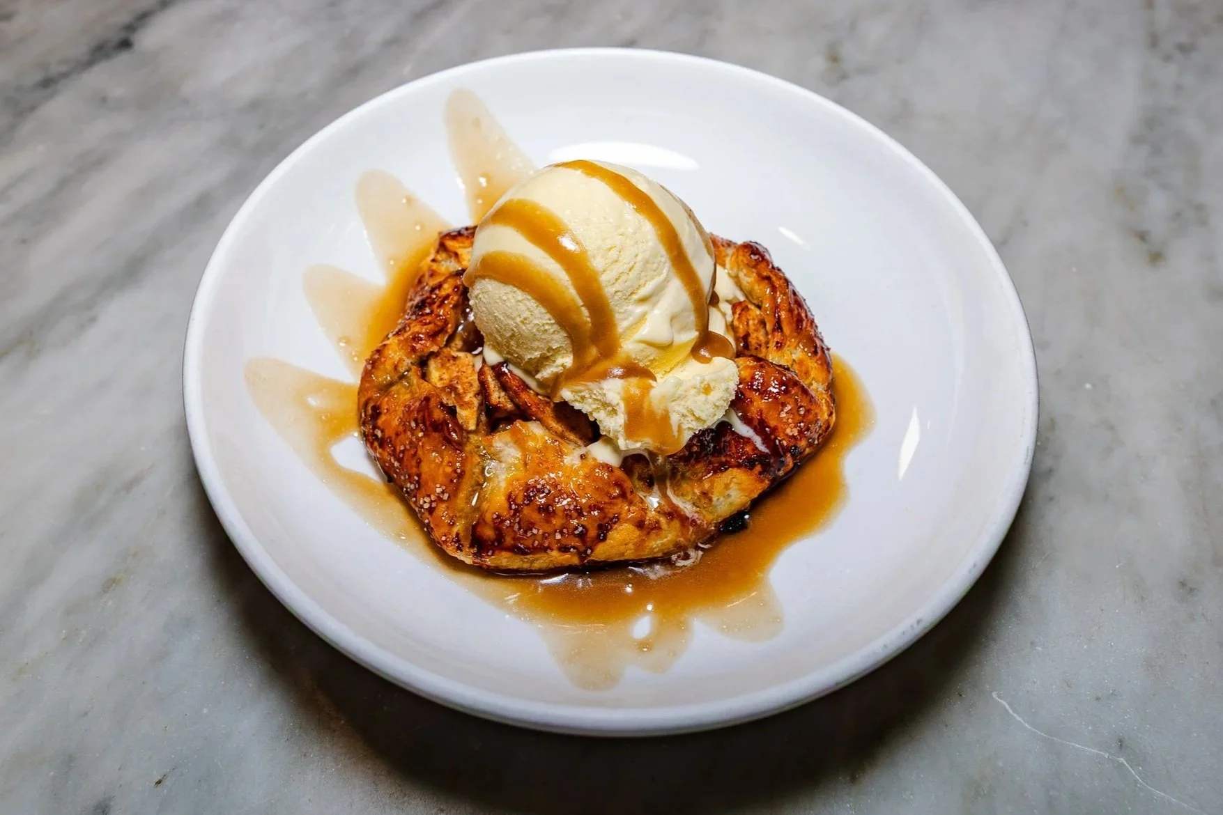 A dessert with a heart-shaped piece of baked pastry topped with a scoop of vanilla ice cream and caramel sauce, served on a white plate.