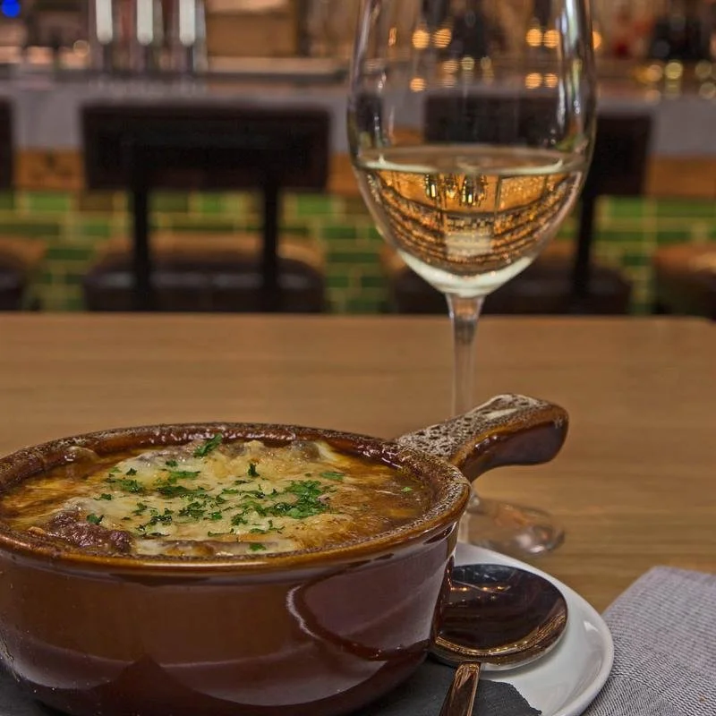 A bowl of baked French onion soup garnished with chopped herbs, served with a glass of white wine on a wooden table in a restaurant setting.