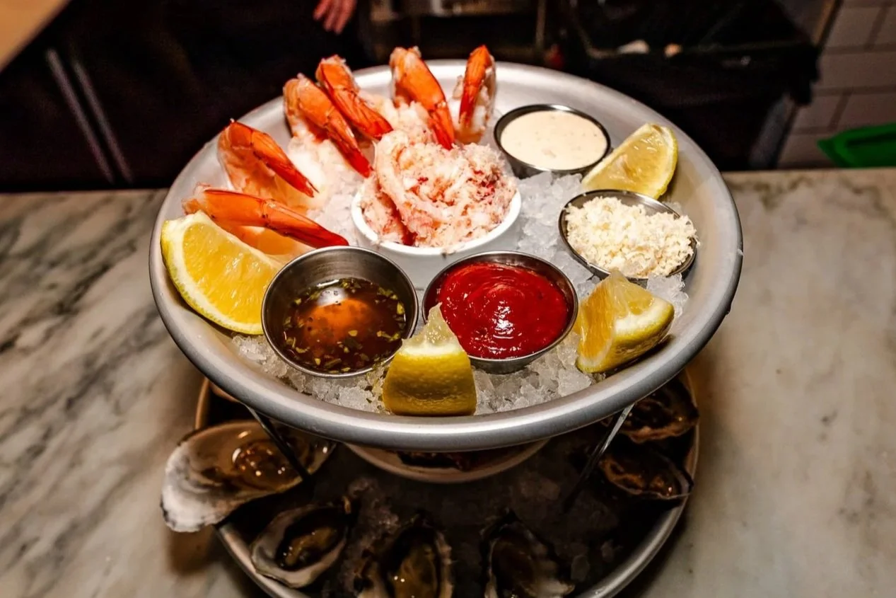 Seafood platter with shrimp, oysters, lemon wedges, cocktail sauce, and dipping sauces on a bed of ice.