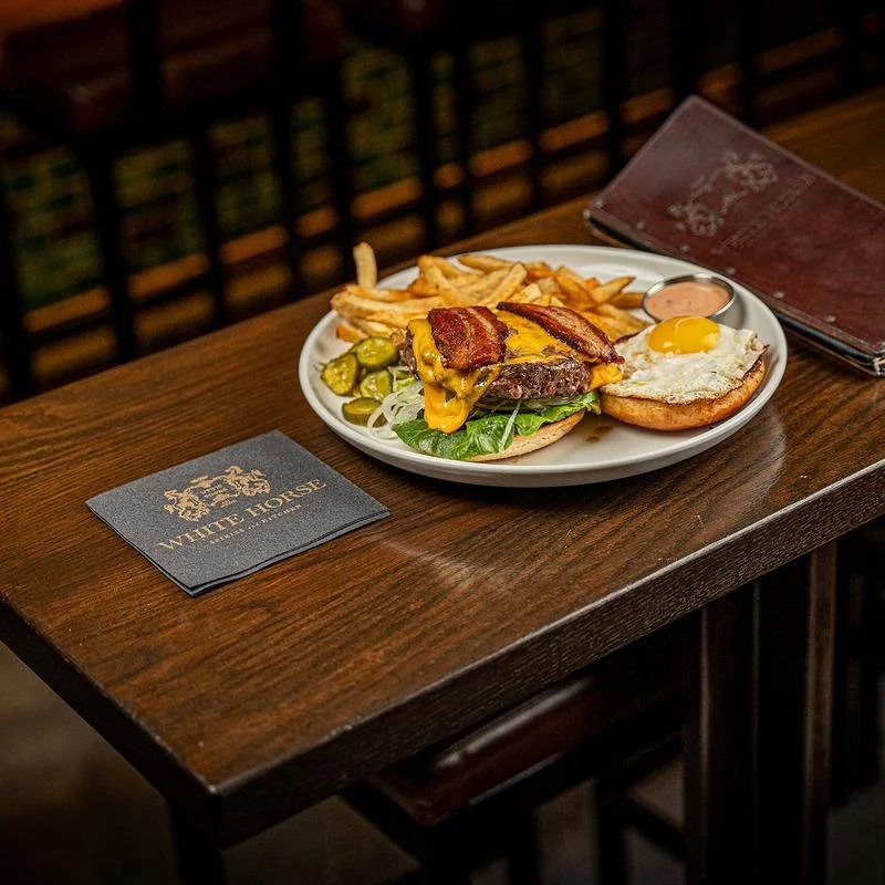 A plate of food with French fries, a burger topped with cheese and bacon, pickles, and a fried egg. There is also a small container of dipping sauce on the plate. The plate is on a wooden table with a menu and a black napkin that reads 'White Horse'.