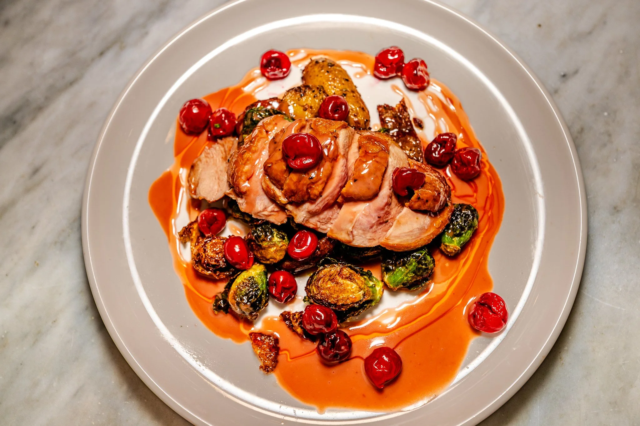 Sliced roasted chicken breast on a bed of charred Brussels sprouts, garnished with red cherries, with a drizzle of orange-colored sauce on a white plate.