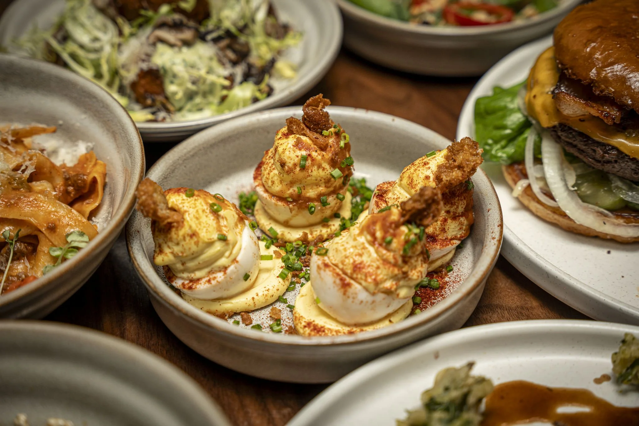 A bowl of deviled eggs topped with chives and paprika surrounded by various bowls of food including a burger with lettuce, onion, cheddar cheese, and bacon and salads.