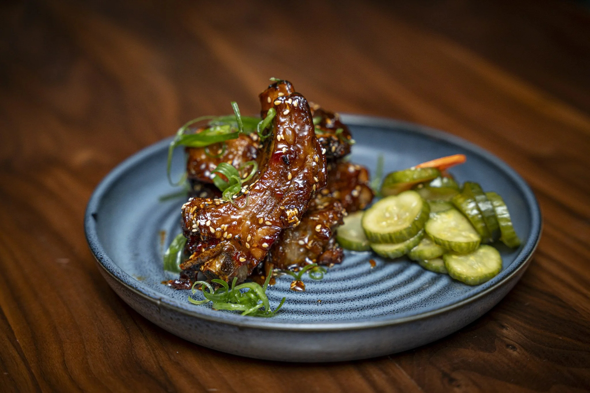 A blue plate with glazed chicken wings garnished with green scallions and sesame seeds, accompanied by sliced cucumbers.