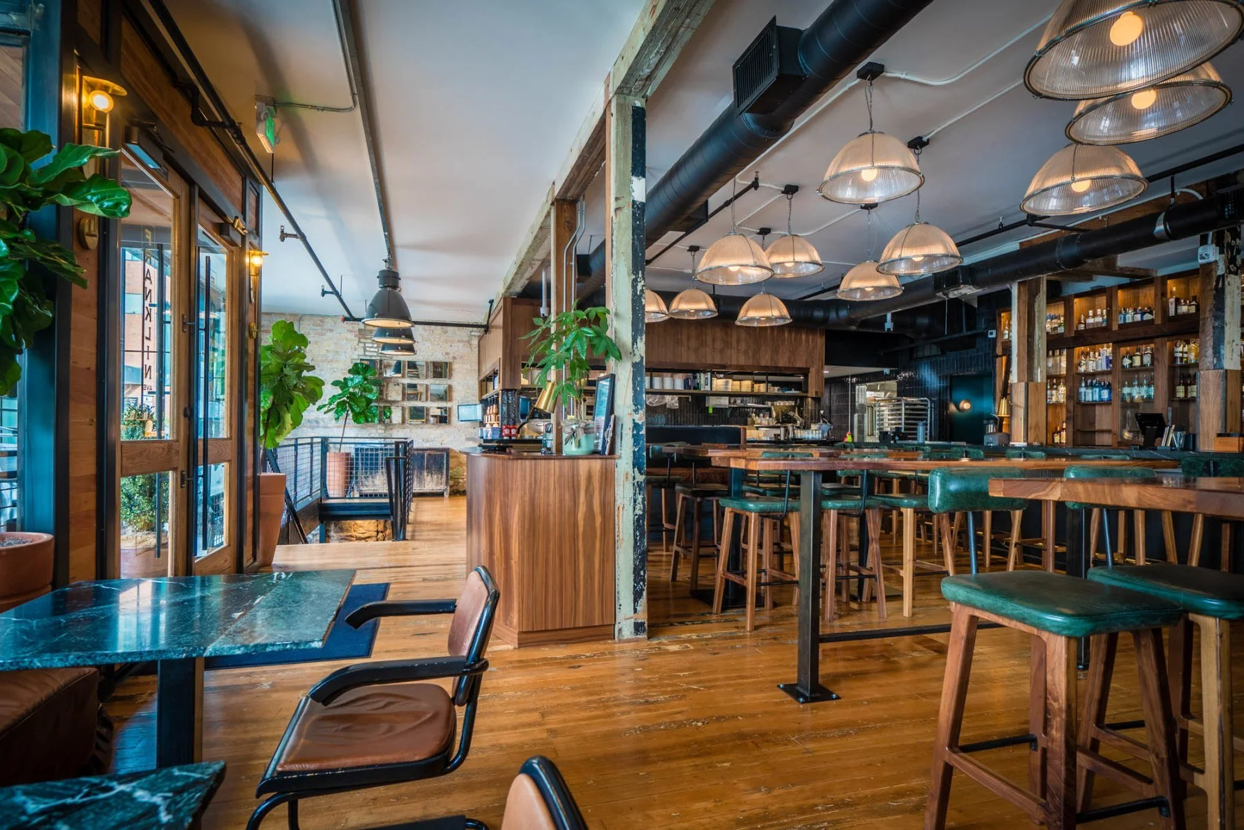 Interior of a cozy restaurant with wooden flooring, green chairs, bar stools, and warm lighting fixtures.