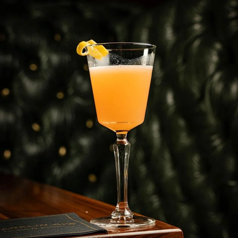 A pink cocktail with a lemon peel garnish in a tall, elegant glass on a wooden surface, with a dark, blurred background.