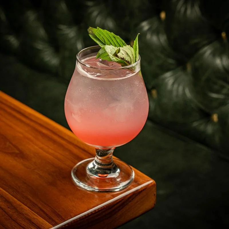 A pink cocktail garnished with fresh mint leaves, served in a stemmed glass on a wooden surface.