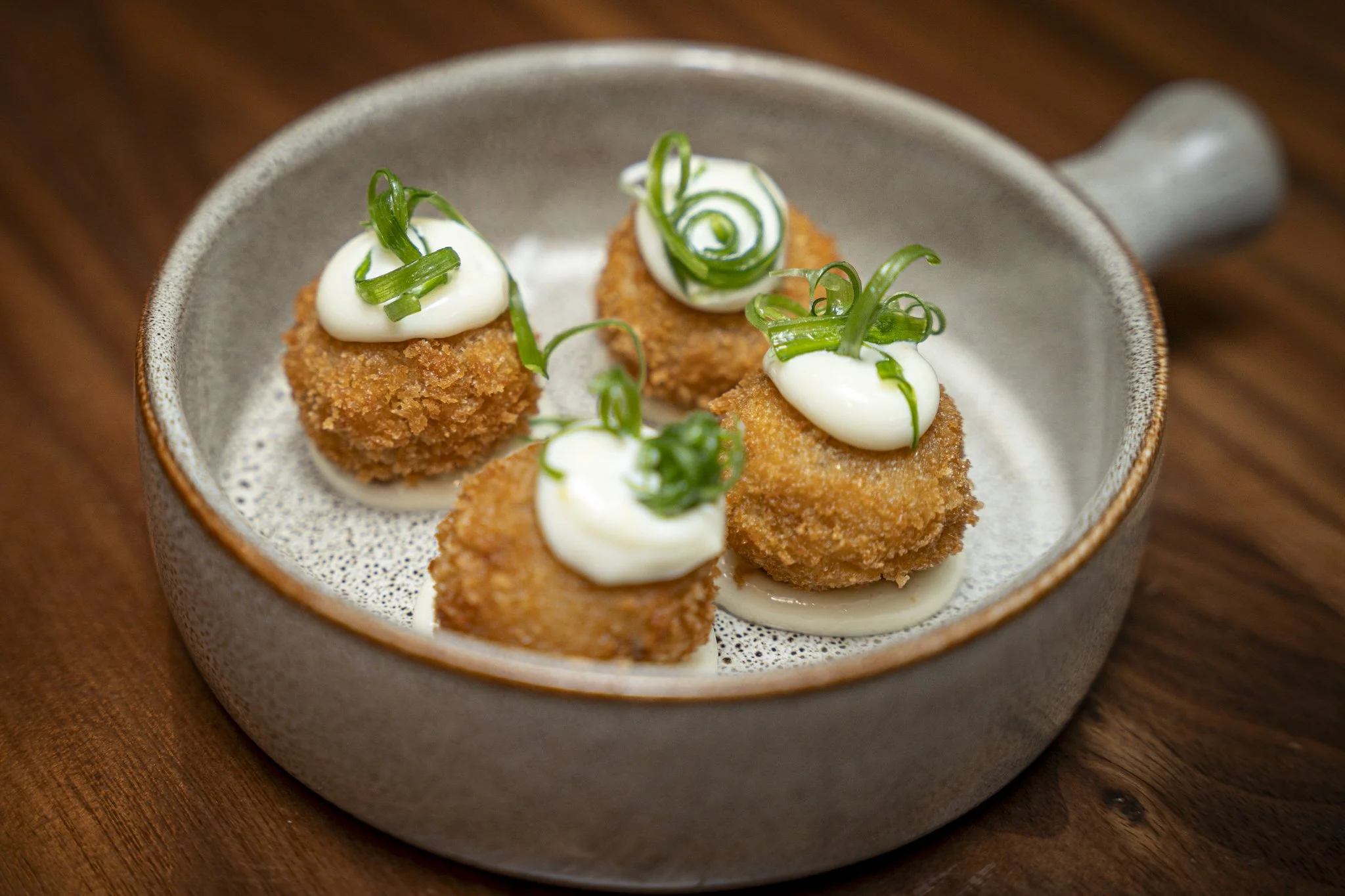 Four breaded and fried appetizers topped with white sauce and garnished with green onion curls in a ceramic dish.
