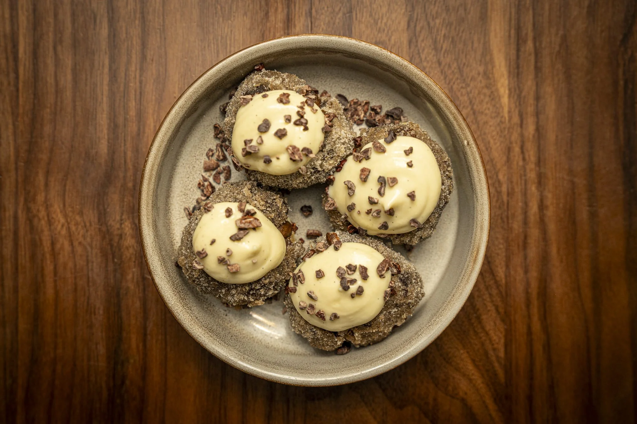 Four chocolate-dipped cookies topped with white chocolate and sprinkled with cocoa nibs, served on a beige ceramic plate on a wooden table.