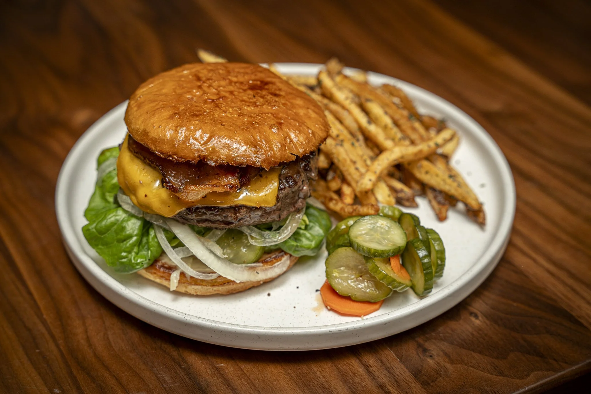 A cheeseburger with lettuce, onion, tomato, bacon, and melted cheese, served with seasoned French fries and pickles on a white plate.