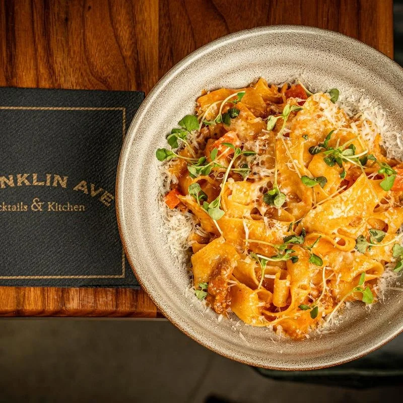 A ceramic bowl of pasta with shredded cheese, fresh herbs, and a tomato-based sauce on a wooden table.