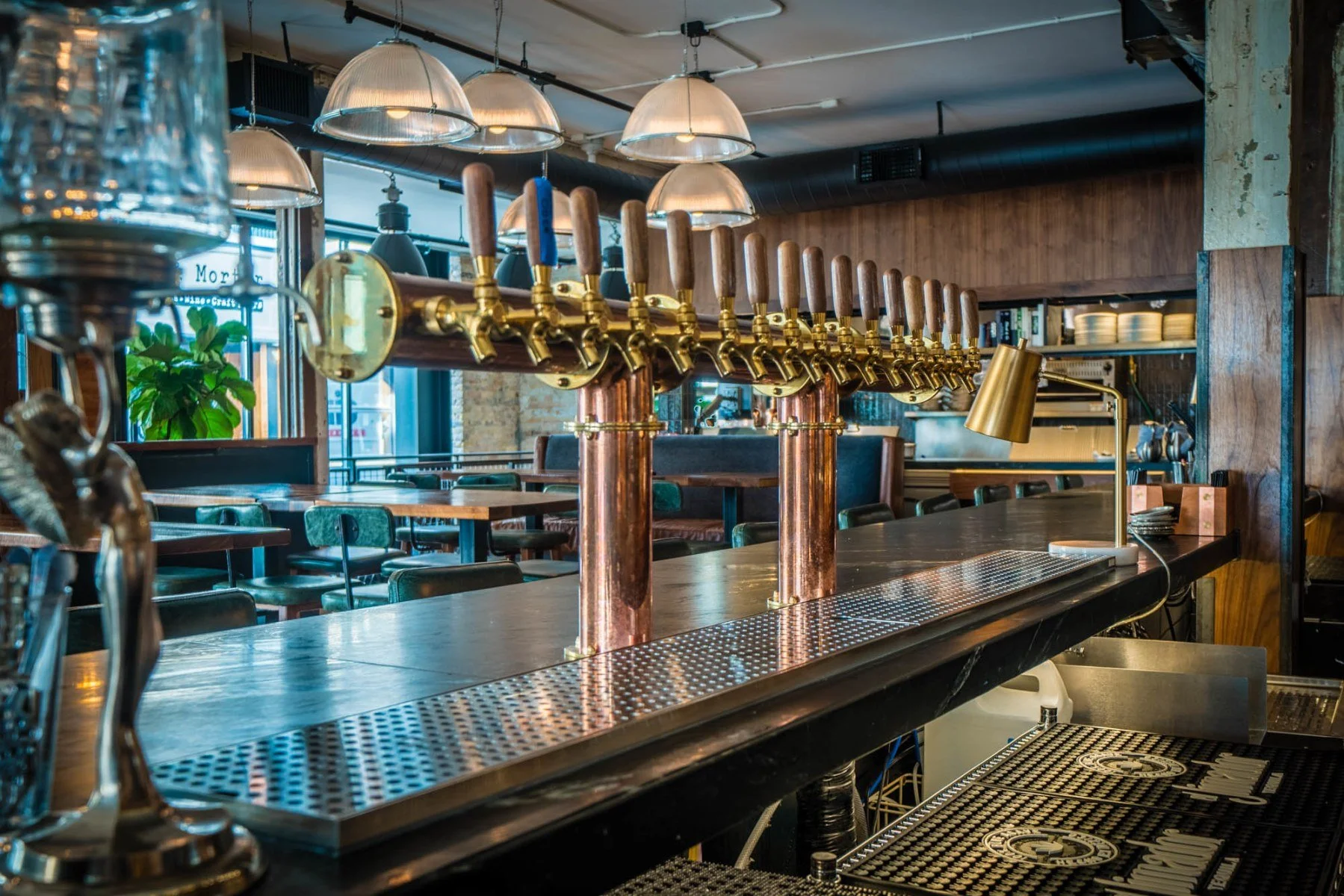 An empty bar with a row of beer taps, a tap handle for a beer or soda, and a glass water dispenser in a modern restaurant with hanging lights, tables, and chairs in the background.