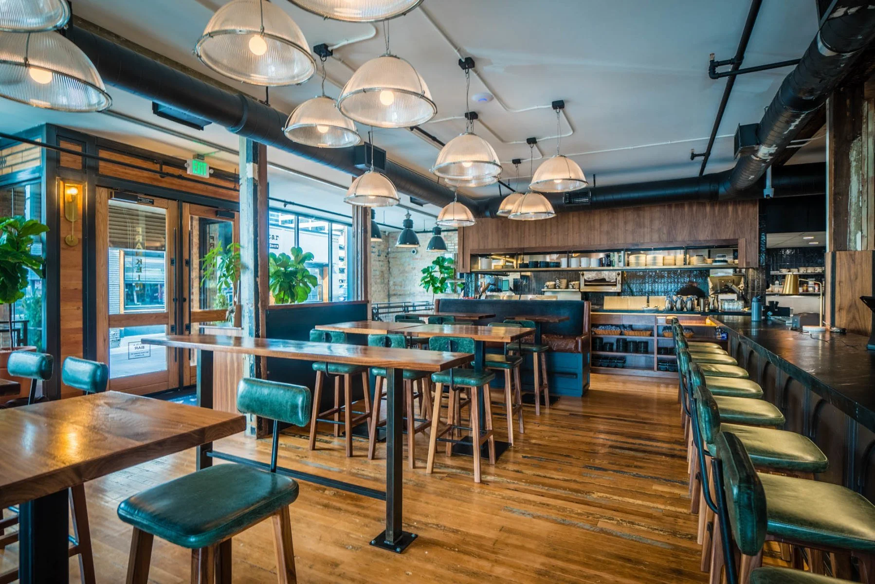 Interior of a modern restaurant or cafe with wooden tables and green chairs, pendant lighting, large windows with plants outside, and an open kitchen area.