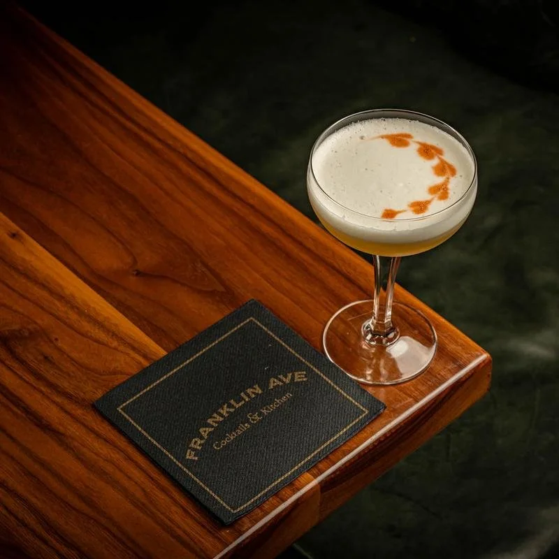 A cocktail glass with a light-colored drink and a circular design made of orange foam on top, placed on a polished wooden bar next to a black napkin that reads 'Franklin Ave Cocktails & More'.
