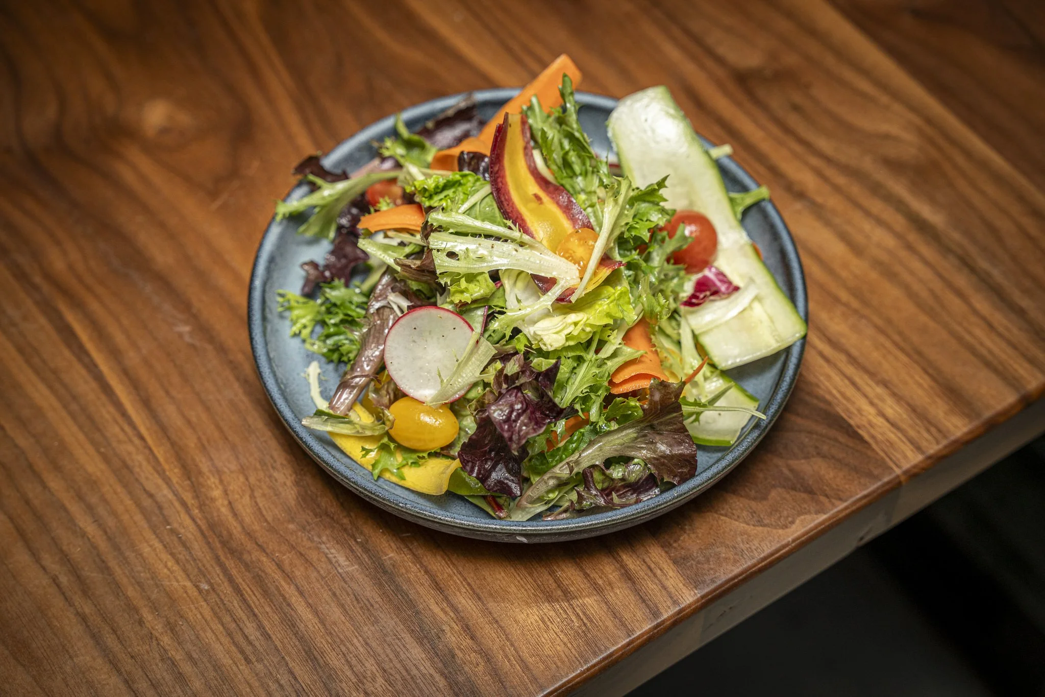 A colorful mixed salad with various vegetables on a blue plate placed on a wooden table.
