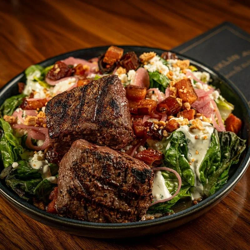 A salad with grilled steak, bacon, lettuce, cheese crumbles, and dressing in a black bowl on a wooden table.