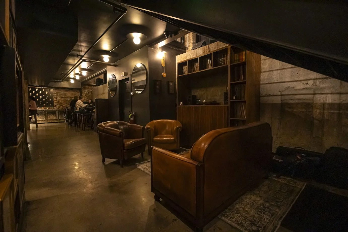 A cozy, dimly lit lounge area with leather armchairs, a wooden bookshelf, and walls decorated with mirrors and vintage decor. In the background, there are people sitting at tables.