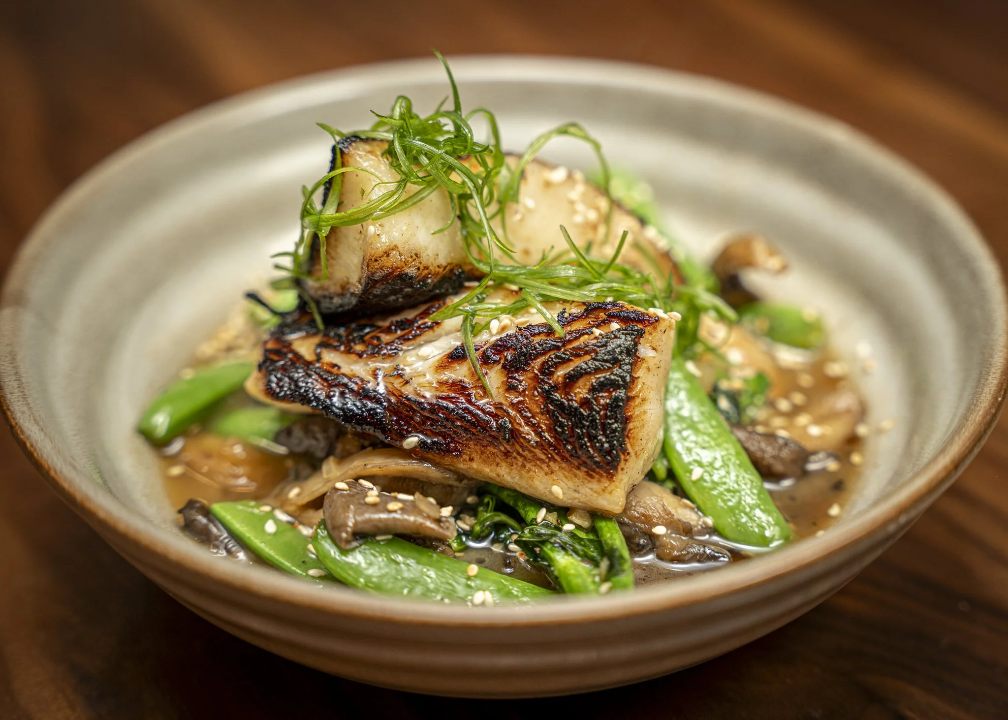 Close-up of a cooked fish fillet with char marks, garnished with green onions, sesame seeds, served with peppers and sauce in a ceramic bowl.