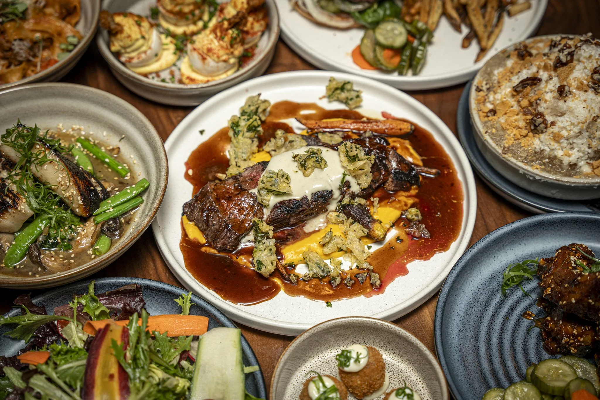A variety of Korean dishes on a table, including a plate of bulgogi with sauce and toppings, a bowl of salad, a plate with cooked fish and vegetables, a bowl of rice, and several side dishes.