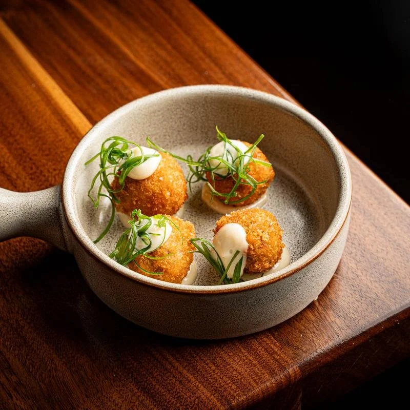 Fried appetizer balls garnished with microgreens and a dollop of sauce in a ceramic dish on a wooden table.
