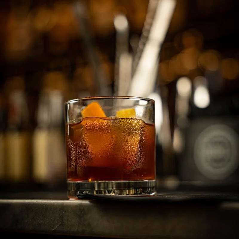 A glass of amber-colored cocktail with ice cubes and orange peel, placed on a dark bar counter with a blurred bar background.