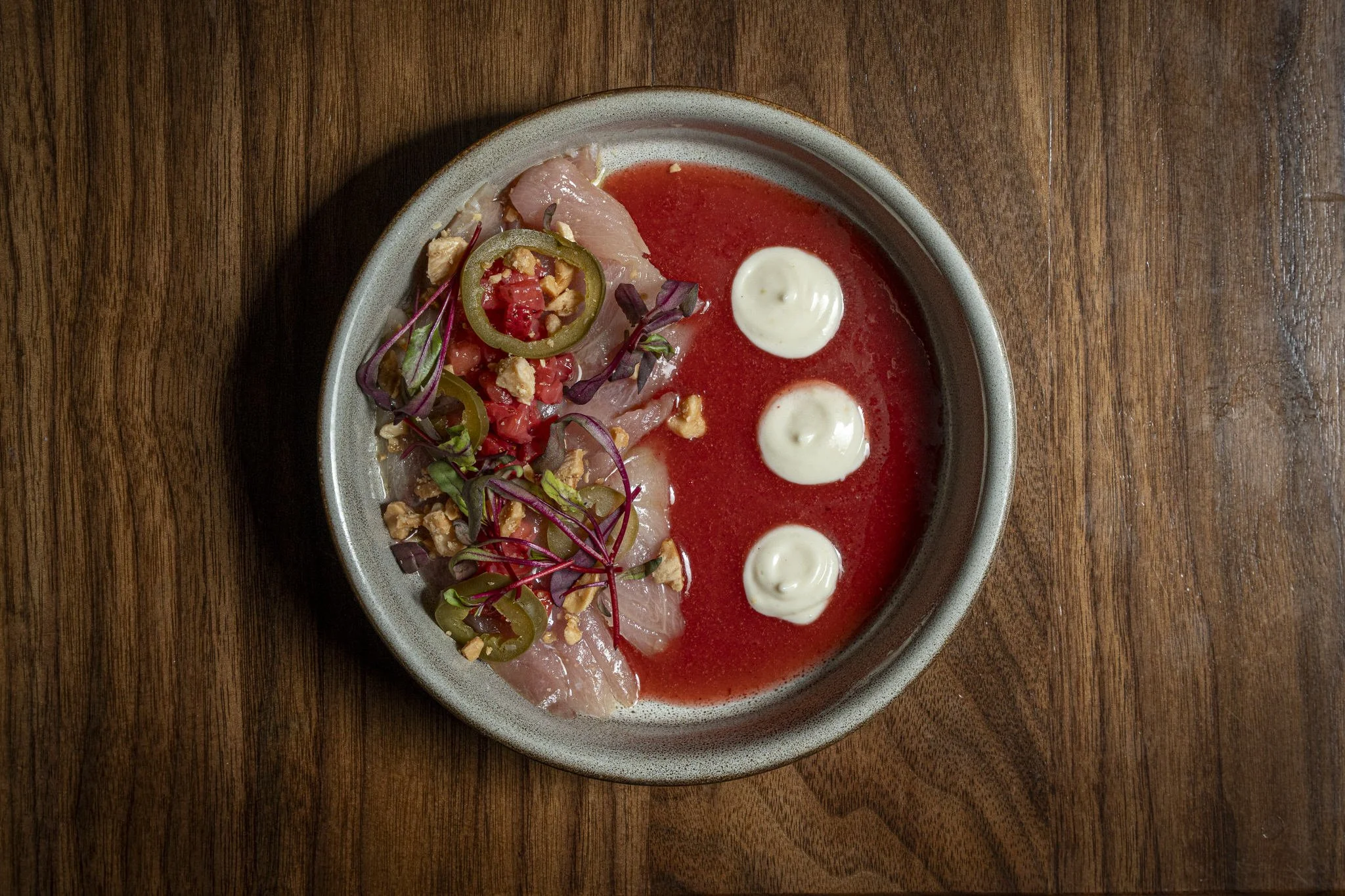 A bowl of raw fish sashimi with sliced jalapeños, microgreens, and chopped nuts on the side, served with a side of red sauce, on a wooden table.