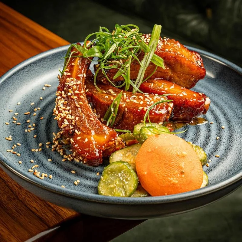 Barbecue ribs garnished with greens, sesame seeds, and served with sliced pickles and carrots on a blue plate.