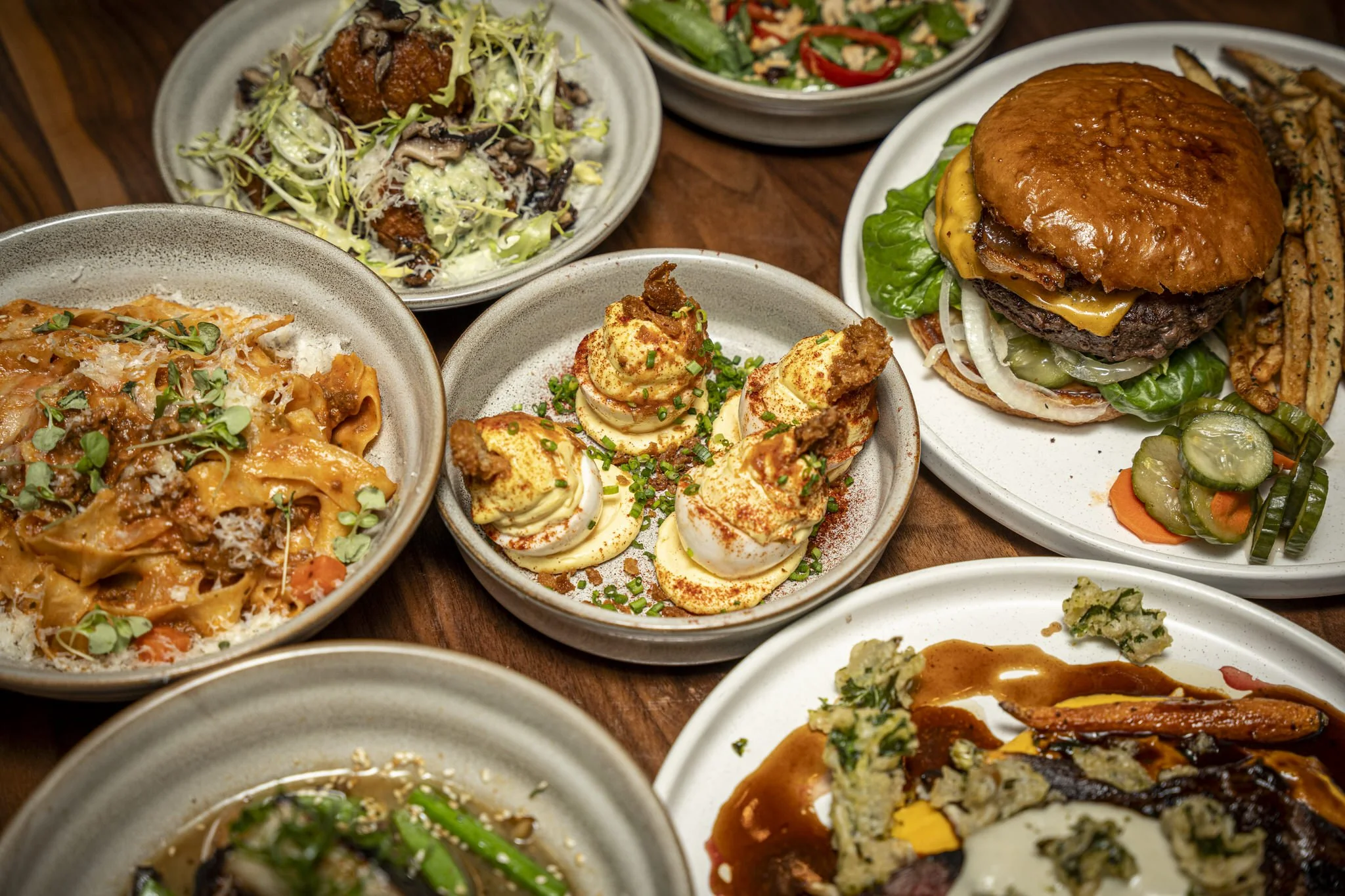 Assorted dishes including a burger with lettuce, tomato, onions, and cheddar cheese, a plate of deviled eggs sprinkled with paprika and chives, a salad with mixed greens, chopped vegetables, and sliced red chili, a pasta with tomato sauce and grated cheese, and other side dishes on a wooden table.