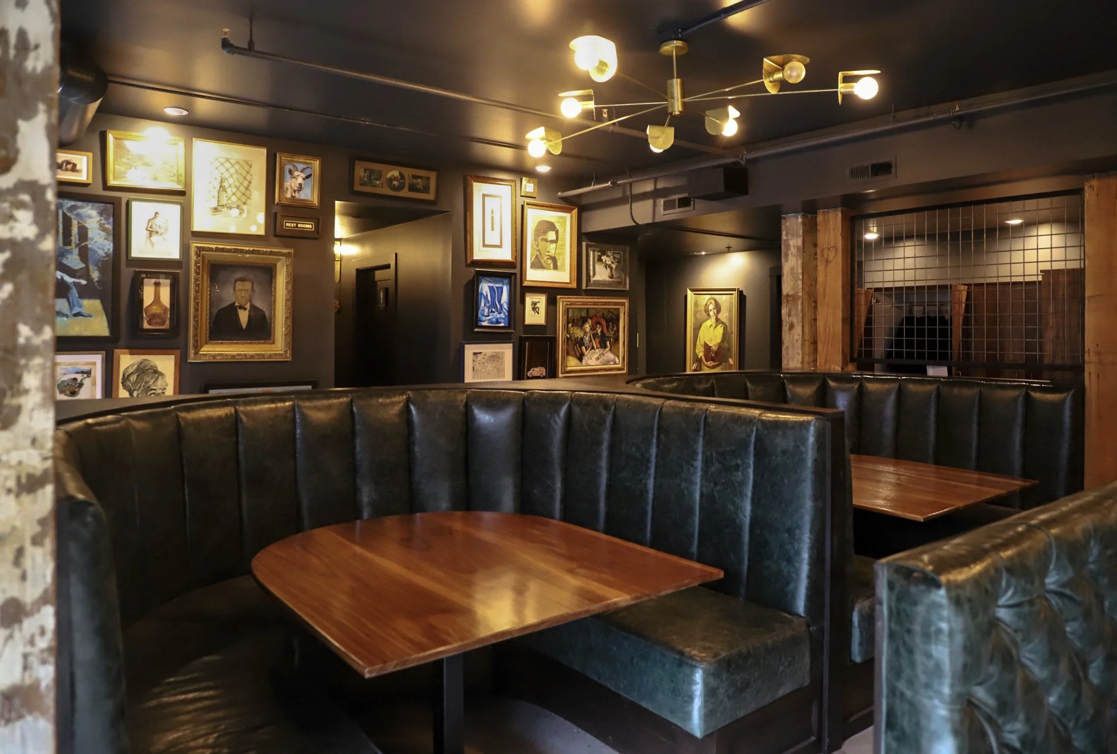 Interior of a cozy restaurant or cafe with dark walls decorated with framed art and photographs, leather booth seating with wooden tables, and warm lighting from a modern chandelier.