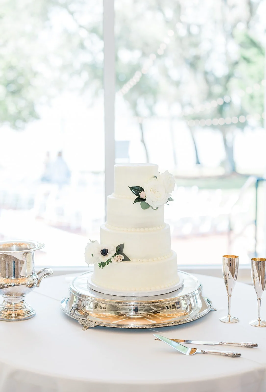 A four-tier white wedding cake with floral decorations on a silver cake stand at a reception table, with silver serving utensils and champagne glasses nearby.