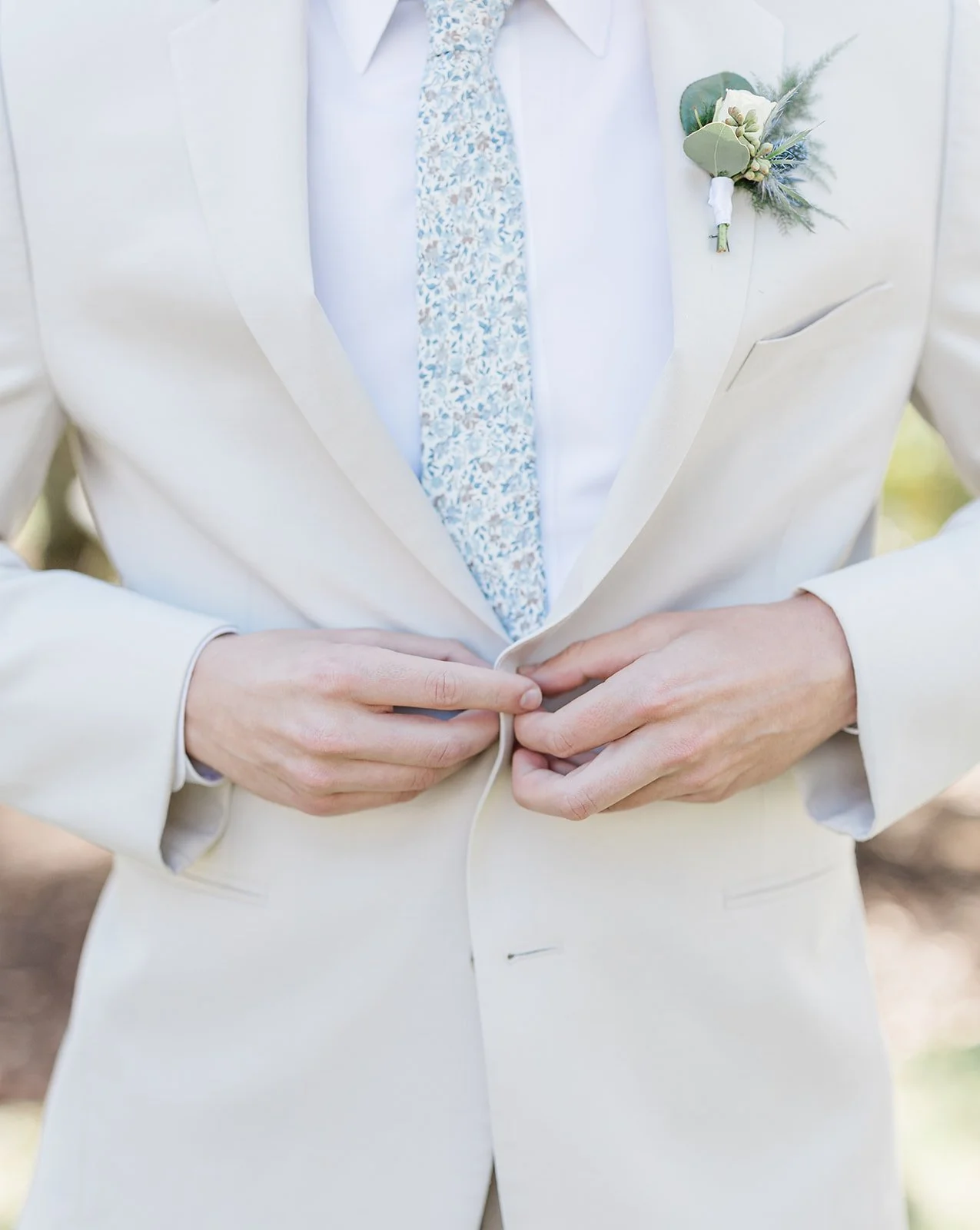 Close-up of a man in a beige suit buttoning his jacket, wearing a light-colored tie with a floral pattern, and a boutonniere with green leaves and small flowers on his left lapel.
