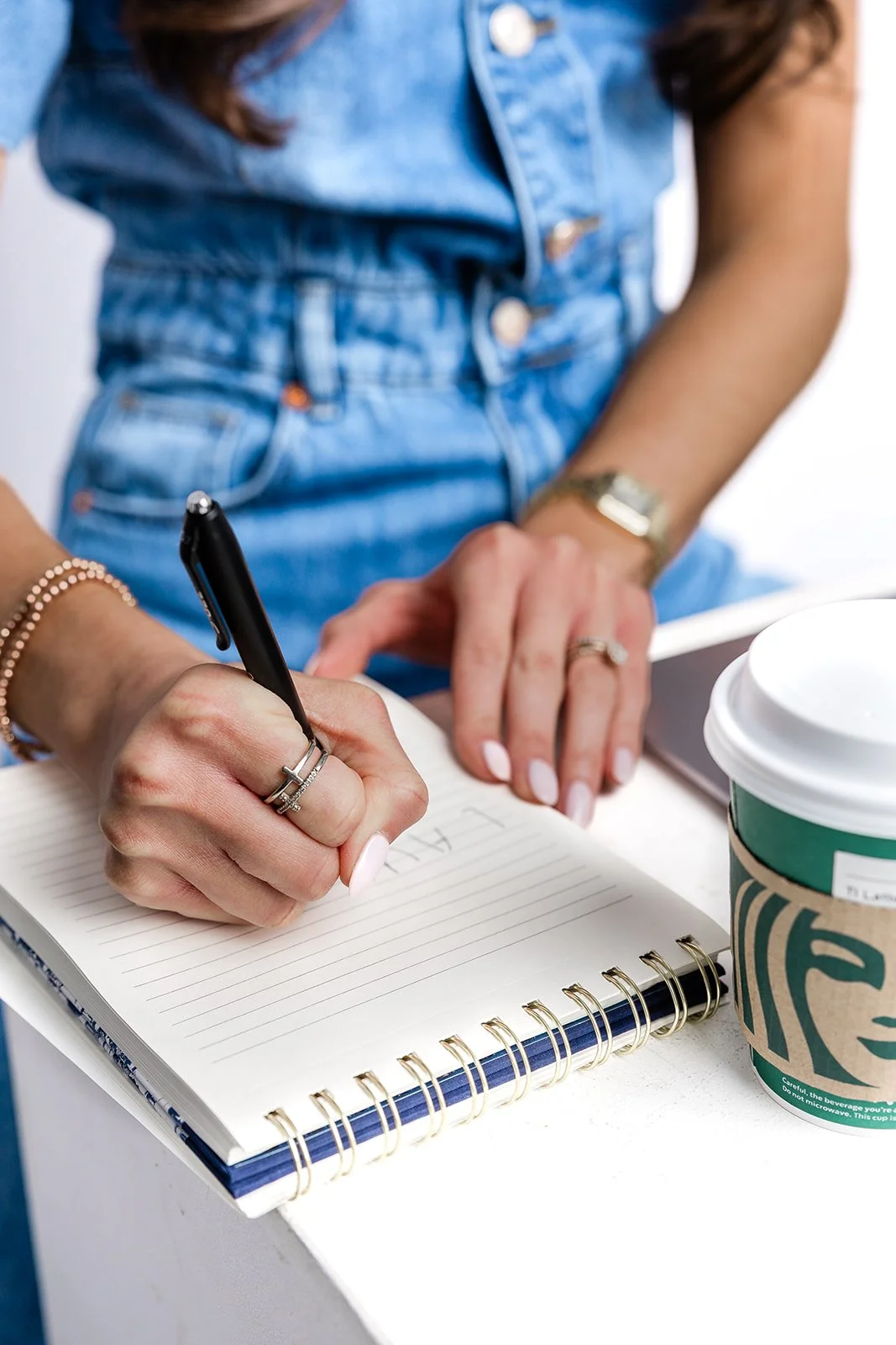A woman wearing a denim jumpsuit and rings is writing in a spiral notebook with a black pen on a white table. There is a Starbucks cup with a green sleeve on the table.