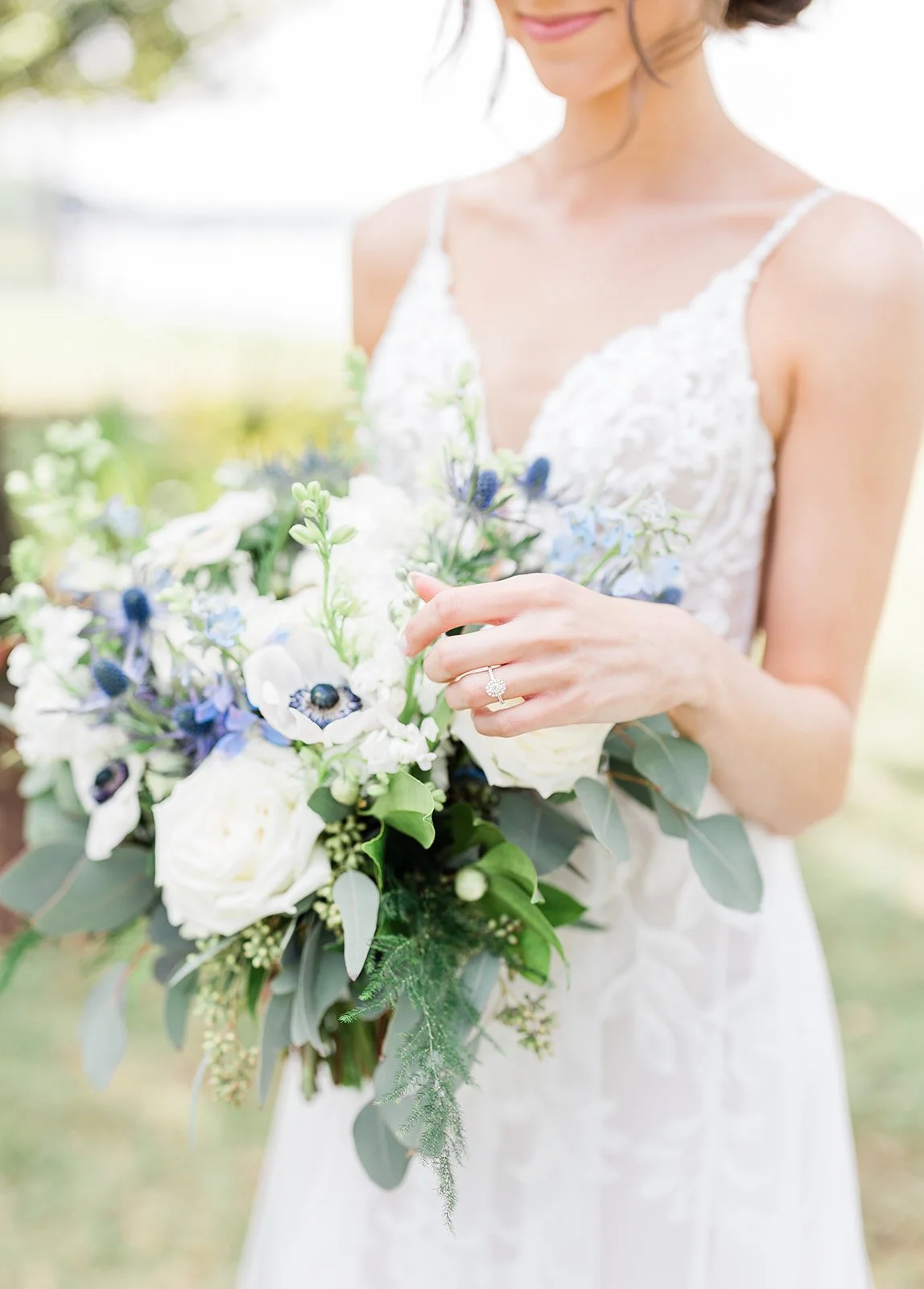 Close-up of a bride holding a bouquet of white and blue flowers, wearing a wedding dress and an engagement ring, outdoors with a soft focus background.
