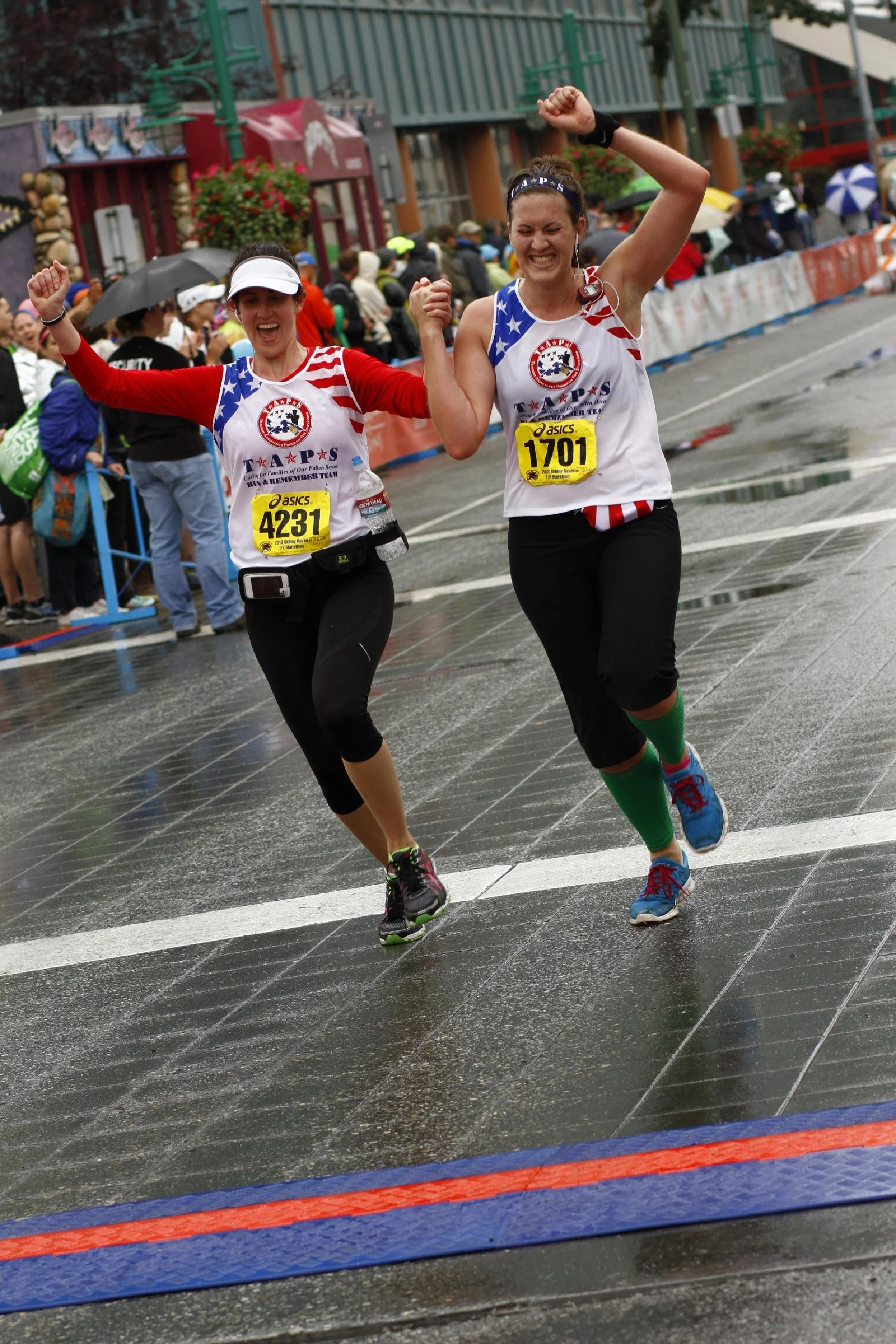 Two women celebrating as they finish a race, crossing the finish line on a rainy day, wearing race bibs and athletic gear.
